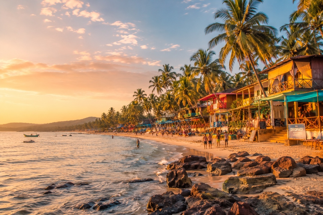 A palm-fringed Goa beach at sunset, with wooden beach shacks lining the shore